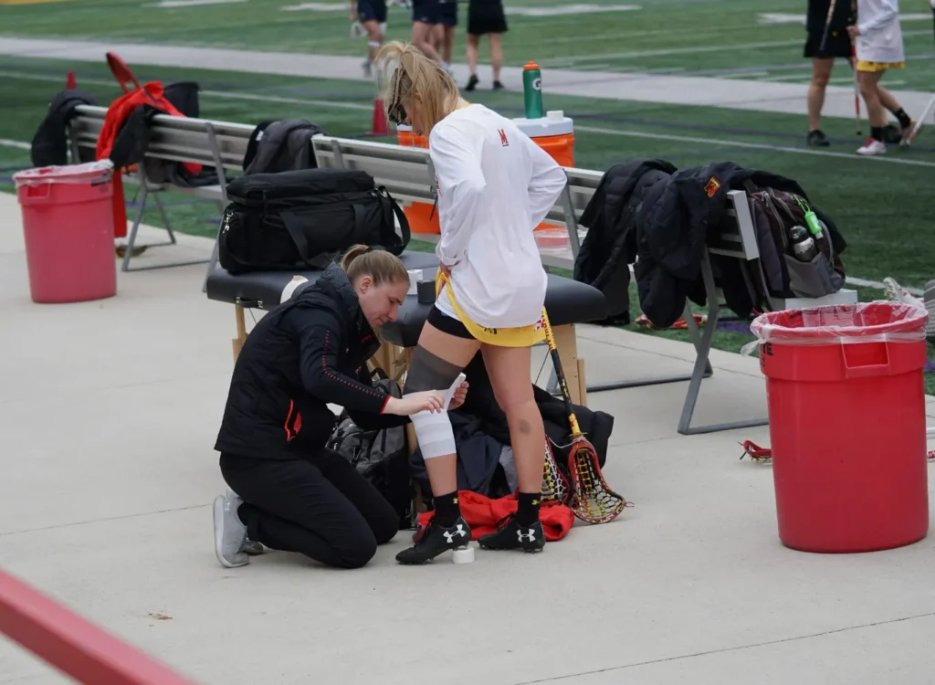 Athletic trainer wrapping a lacrosse player's knee on the sidelines during a game.
