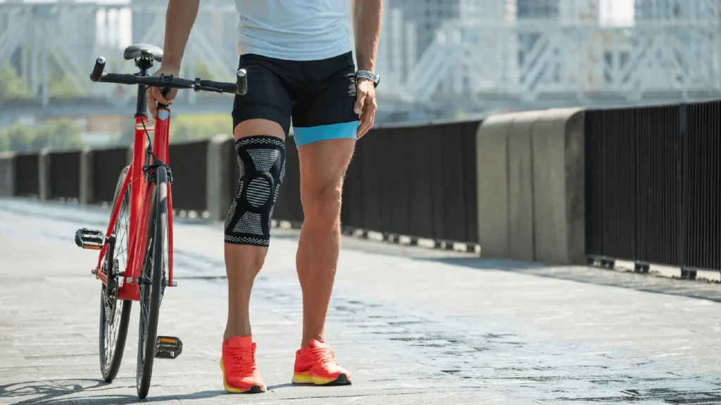 A person wearing a knee brace on the left leg stands next to a red bicycle on a paved pathway with a bridge in the background.