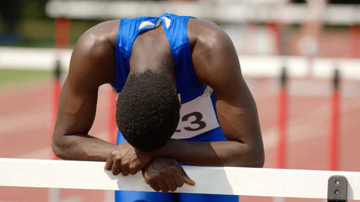 A male track athlete in a blue uniform leans over a hurdle on a track, head resting on his hands, appearing exhausted or disappointed.