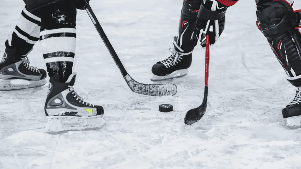 Photograph showing a close-up of two ice hockey players' legs and sticks competing for a puck on an ice rink. Players are avoiding winter hockey injuries.