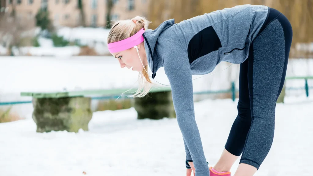 A person in a grey jacket and pink headband stretching in the snow before exercising to prevent joint pain